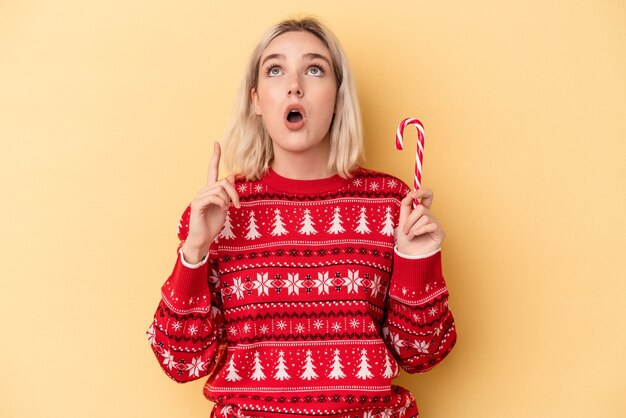 Jovem mulher caucasiana, segurando uma vara de Natal isolada em um fundo amarelo, apontando para cima com a boca aberta.