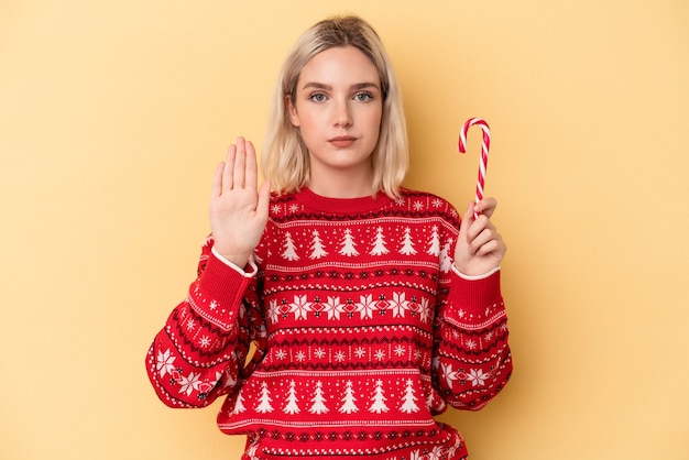 Jovem mulher caucasiana, segurando uma vara de Natal isolada em um fundo amarelo, de pé com a mão estendida, mostrando o sinal de stop, impedindo-o.