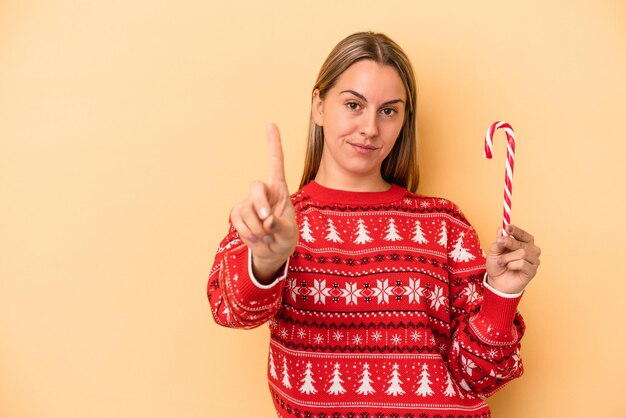 Jovem mulher caucasiana, segurando uma vara de Natal isolada em um fundo amarelo, mostrando o número um com o dedo.