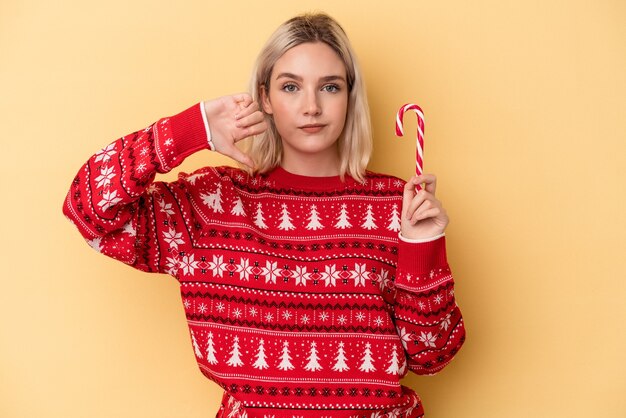 Jovem mulher caucasiana segurando uma vara de Natal isolada em um fundo amarelo, mostrando um gesto de antipatia, polegares para baixo. Conceito de desacordo.