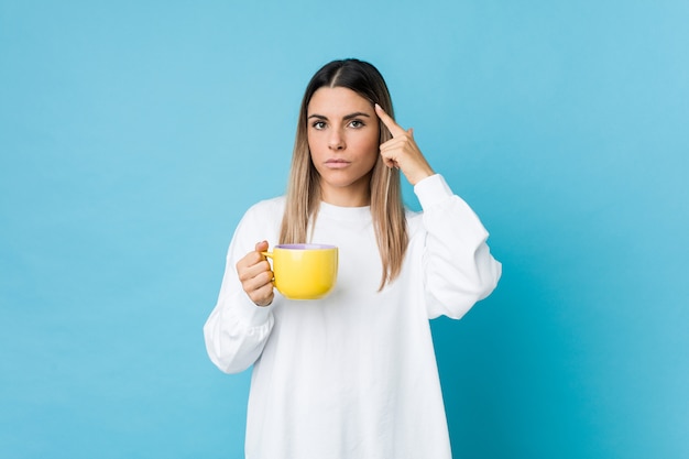 Jovem mulher caucasiana, segurando uma xícara de café, apontando seu templo com o dedo, pensando, focado em uma tarefa.