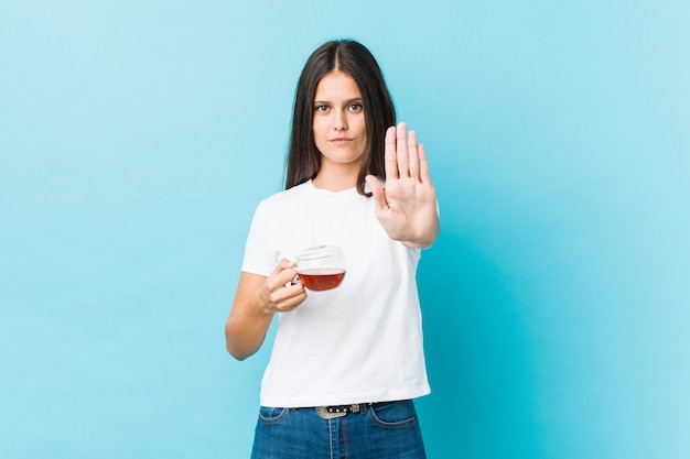 Jovem mulher caucasiana, segurando uma xícara de chá em pé com a mão estendida, mostrando o sinal de stop, impedindo-o.