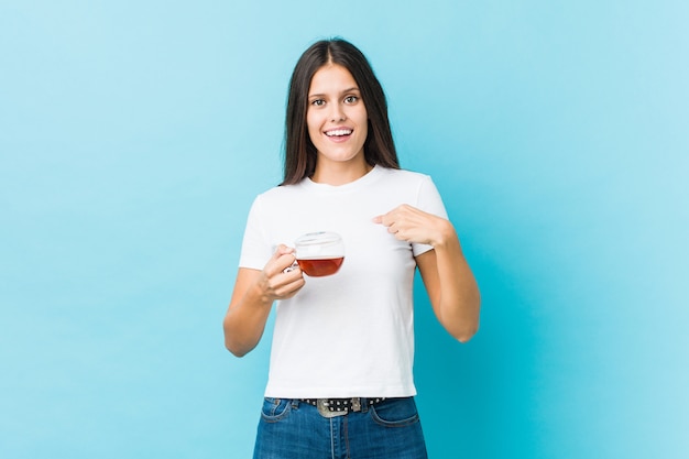 Jovem mulher caucasiana, segurando uma xícara de chá surpreendeu apontando para si mesmo, sorrindo amplamente.