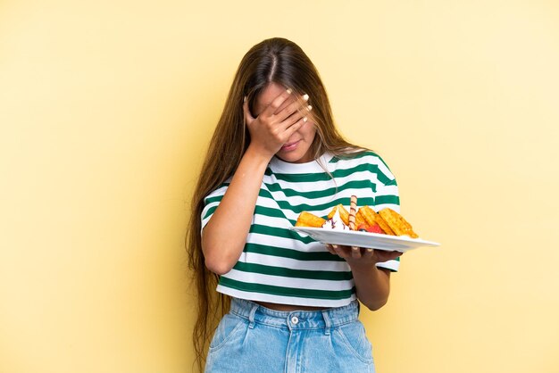 Foto jovem mulher caucasiana segurando waffles isolados em fundo amarelo com expressão cansada e doente