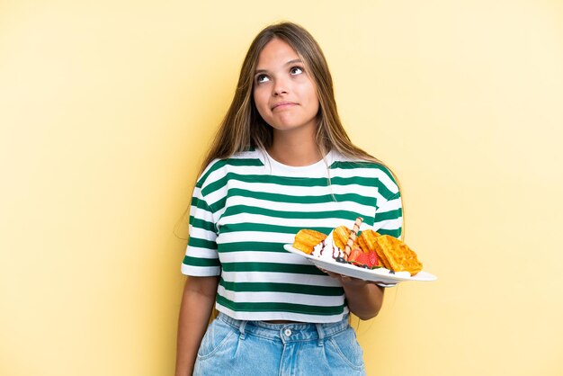 Jovem mulher caucasiana segurando waffles isolados em fundo amarelo e olhando para cima