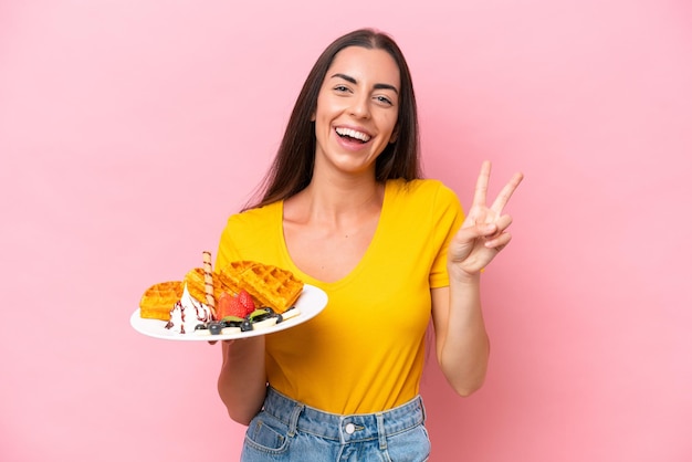 Foto jovem mulher caucasiana segurando waffles isolados em fundo rosa sorrindo e mostrando sinal de vitória