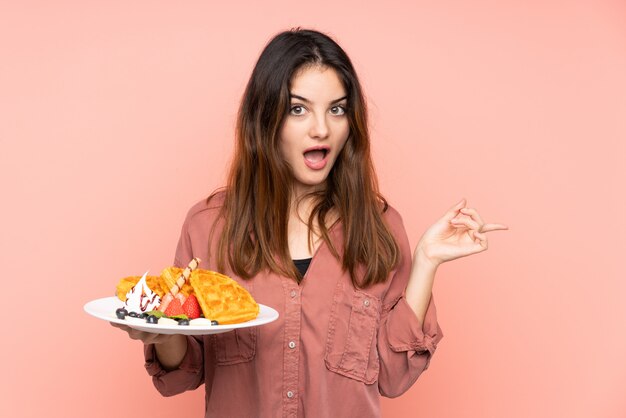 Jovem mulher caucasiana segurando waffles isolados na parede rosa surpreendeu e apontando o dedo para o lado