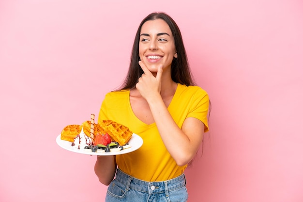 Jovem mulher caucasiana segurando waffles isolados no fundo rosa, olhando para o lado e sorrindo