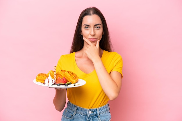 Jovem mulher caucasiana segurando waffles isolados no pensamento de fundo rosa