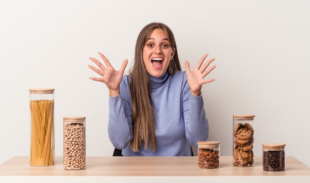 Jovem mulher caucasiana, sentada à mesa com a panela de comida isolada no fundo branco, comemorando uma vitória ou sucesso, ele fica surpreso e chocado.