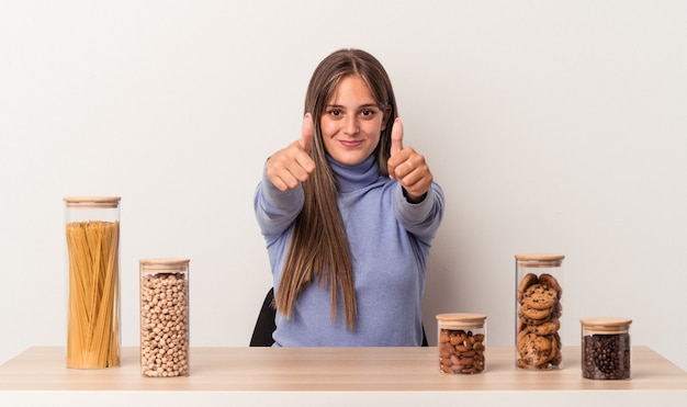 Jovem mulher caucasiana, sentada à mesa com o pote de comida, isolado no fundo branco com polegares para cima, elogios sobre algo, conceito de apoio e respeito.