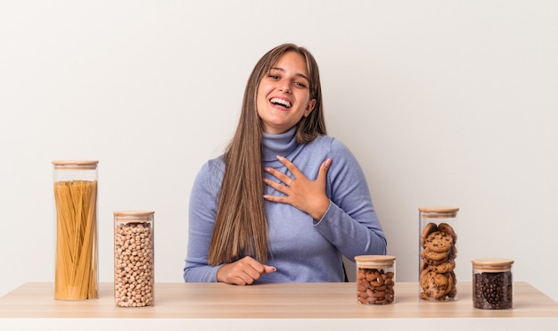 Jovem mulher caucasiana, sentada à mesa com o pote de comida, isolado no fundo branco, ri alto, mantendo a mão no peito.