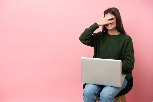 Jovem mulher caucasiana sentada em uma cadeira com seu laptop isolado em um fundo rosa, cobrindo os olhos pelas mãos e sorrindo