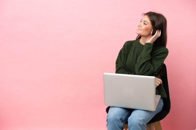 Jovem mulher caucasiana sentada em uma cadeira com seu laptop isolado em um fundo rosa, pensando em uma ideia