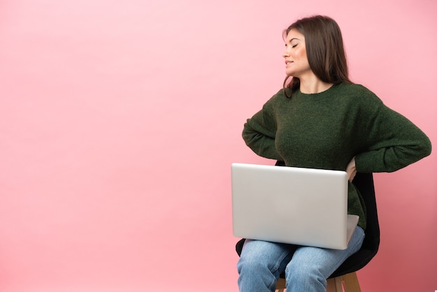 Jovem mulher caucasiana sentada em uma cadeira com seu laptop isolado em um fundo rosa, sofrendo de dor nas costas por ter feito um esforço