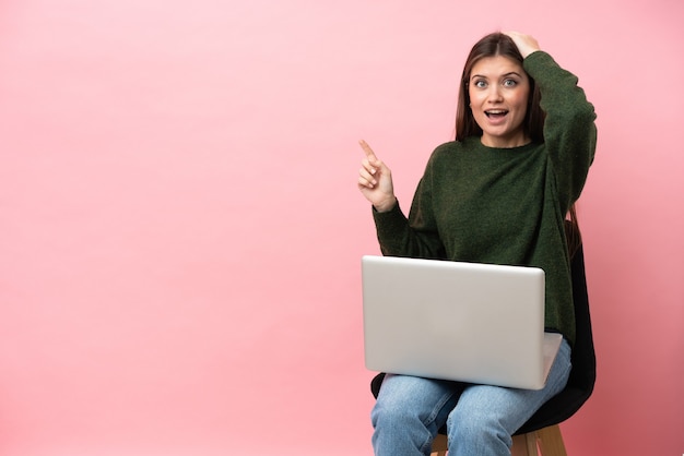 Jovem mulher caucasiana sentada em uma cadeira com seu laptop isolado em um fundo rosa surpresa e apontando o dedo para o lado