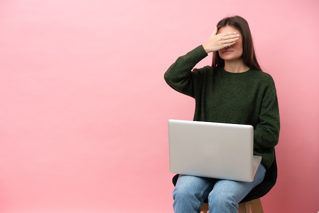 Jovem mulher caucasiana, sentada em uma cadeira com seu laptop isolado no fundo rosa, cobrindo os olhos com as mãos. Não quero ver nada
