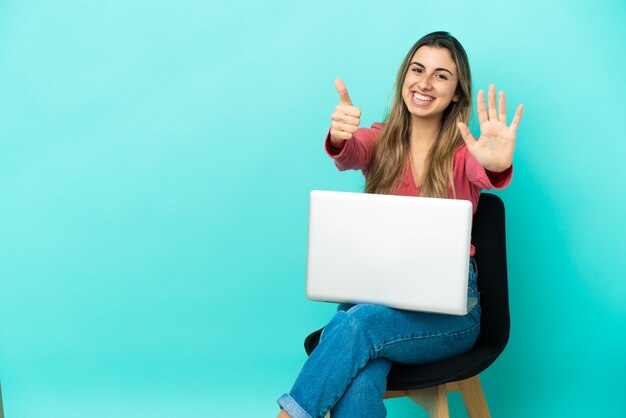 Jovem mulher caucasiana sentada em uma cadeira com seu pc isolado em um fundo azul, contando seis com os dedos