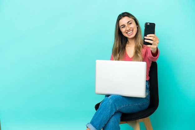 Jovem mulher caucasiana sentada em uma cadeira com seu pc isolado em um fundo azul fazendo uma selfie