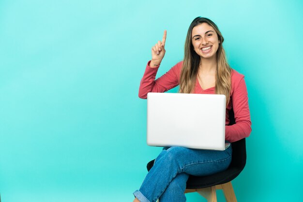 Jovem mulher caucasiana sentada em uma cadeira com seu pc isolado em um fundo azul, mostrando e levantando um dedo em sinal dos melhores