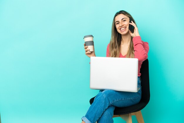 Jovem mulher caucasiana sentada em uma cadeira com seu pc isolado em um fundo azul segurando um café para levar e um celular