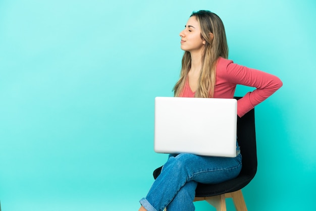 Jovem mulher caucasiana sentada em uma cadeira com seu pc isolado em um fundo azul, sofrendo de dor nas costas por ter feito um esforço