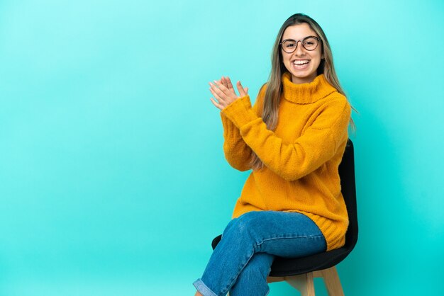 Jovem mulher caucasiana sentada em uma cadeira isolada em um fundo azul, aplaudindo após uma apresentação em uma conferência