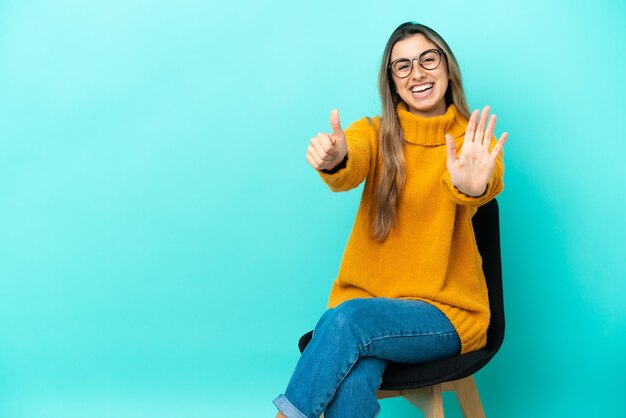 Jovem mulher caucasiana sentada em uma cadeira isolada em um fundo azul contando seis com os dedos