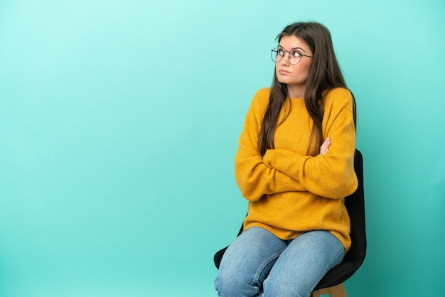 Jovem mulher caucasiana sentada em uma cadeira isolada em um fundo azul fazendo gestos de dúvida enquanto levanta os ombros