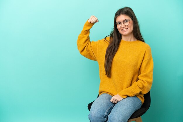 Jovem mulher caucasiana sentada em uma cadeira isolada em um fundo azul fazendo um gesto forte