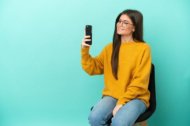 Jovem mulher caucasiana sentada em uma cadeira isolada em um fundo azul fazendo uma selfie