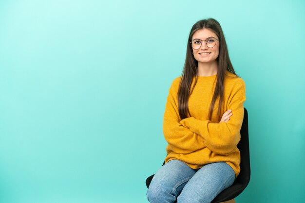 Jovem mulher caucasiana, sentada em uma cadeira isolada em um fundo azul, mantendo os braços cruzados na posição frontal