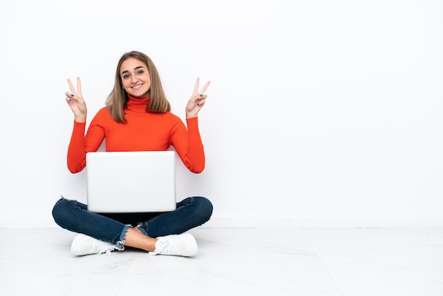 Jovem mulher caucasiana sentada no chão com um laptop mostrando sinal de vitória com as duas mãos