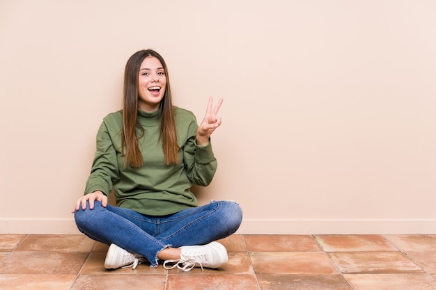 Jovem mulher caucasiana sentada no chão isolada alegre e despreocupada, mostrando um símbolo de paz com os dedos.