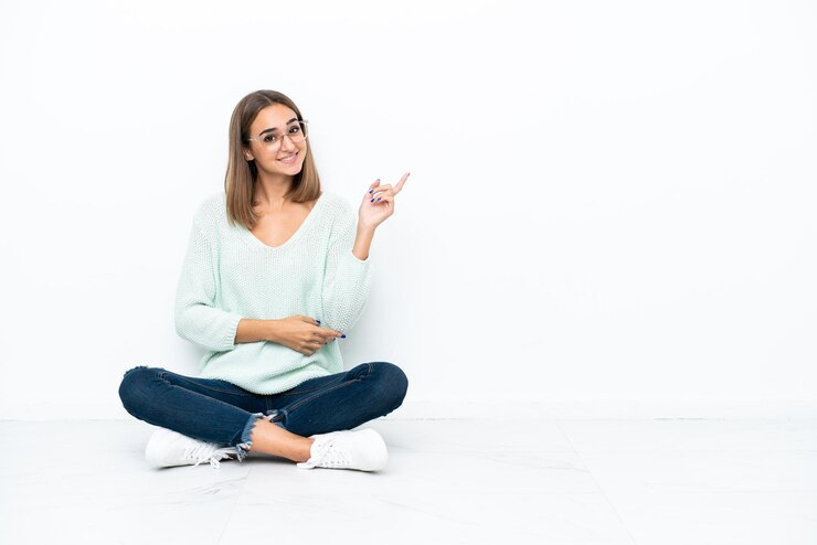 Foto jovem mulher caucasiana sentada no chão isolada no fundo branco apontando o dedo para o lado