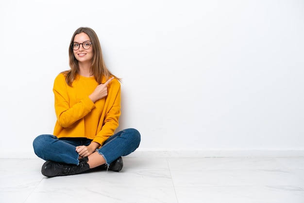 Jovem mulher caucasiana sentada no chão isolada no fundo branco apontando para o lado para apresentar um produto