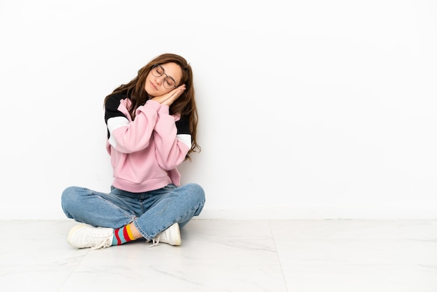 Jovem mulher caucasiana sentada no chão, isolada no fundo branco, fazendo gestos para dormir em expressão dorível