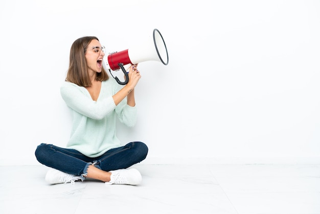 Jovem mulher caucasiana sentada no chão, isolada no fundo branco, gritando através de um megafone