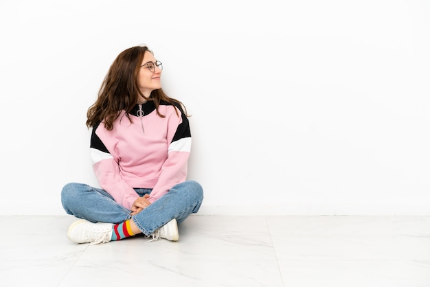 Foto jovem mulher caucasiana sentada no chão, isolada no fundo branco, olhando para o lado