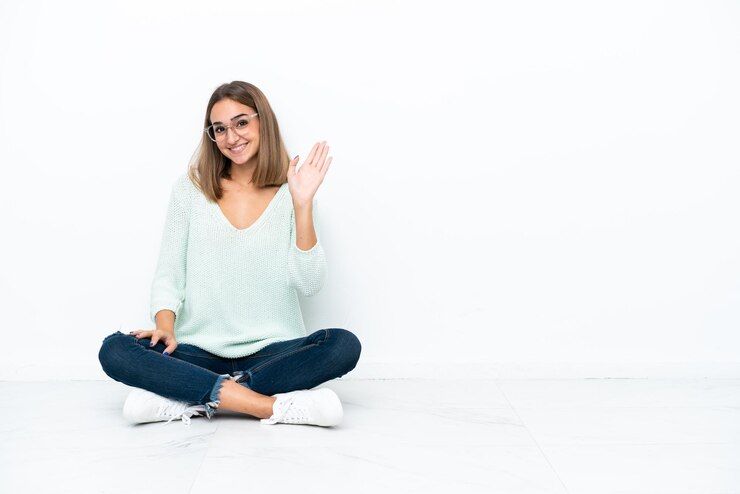 Foto jovem mulher caucasiana sentada no chão, isolada no fundo branco, saudando com a mão com expressão feliz