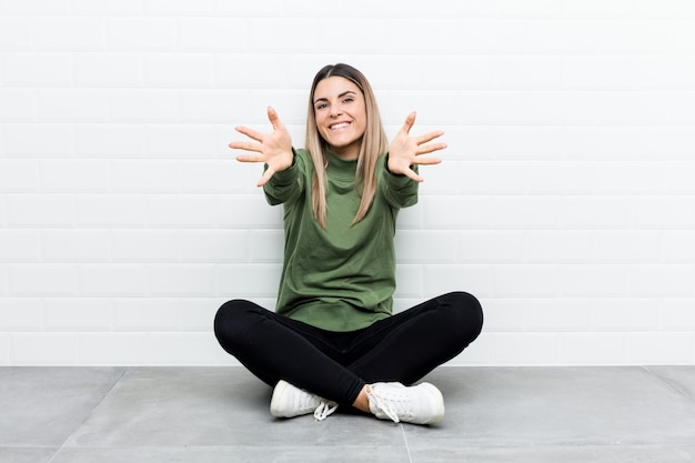 Jovem mulher caucasiana, sentada no chão se sente confiante, dando um abraço para a câmera.