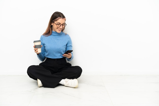 Jovem mulher caucasiana sentada no chão segurando um café para levar e um celular