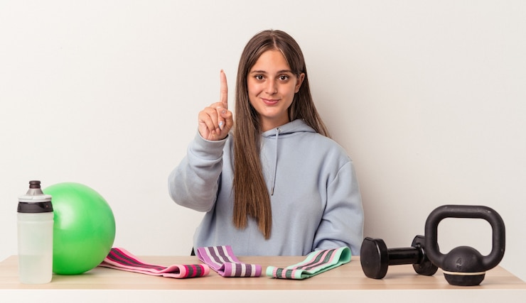 Foto jovem mulher caucasiana, sentado a uma mesa com equipamentos de esporte, isolado no fundo branco, mostrando o número um com o dedo.