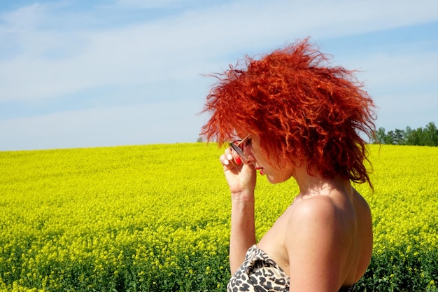 Jovem mulher caucasiana sexy com cabelo vermelho e óculos de sol em fundo de campo de estupro amarelo, dia de verão