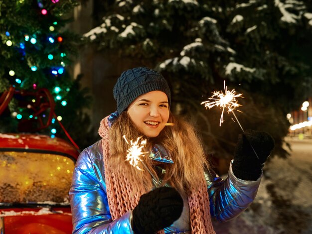 Jovem mulher caucasiana sorridente com cachecol, chapéu, jaqueta, luvas e diamante perto da árvore de natal