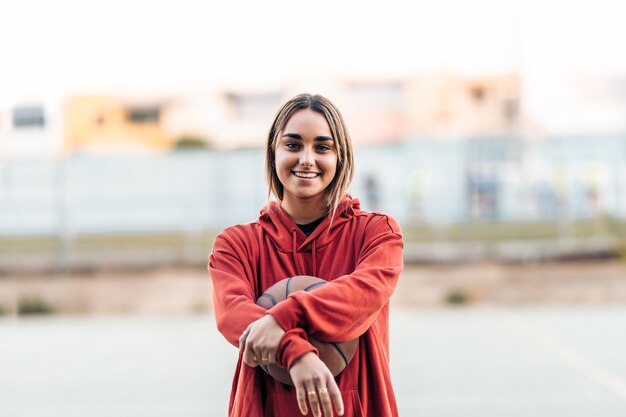 Jovem mulher caucasiana sorridente em roupas esportivas abraçando uma bola de basquete em uma quadra ao ar livre