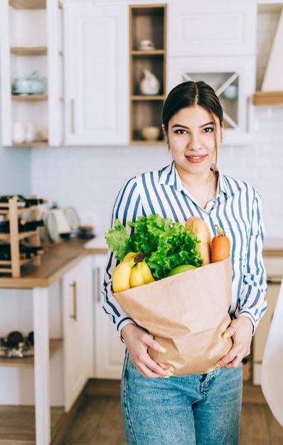 Jovem mulher caucasiana sorridente segura saco de compras eco com legumes frescos e baguete na cozinha moderna.