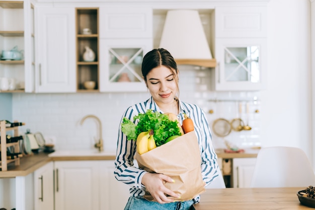 Jovem mulher caucasiana sorridente segura saco de compras eco com legumes frescos e baguete na cozinha moderna.