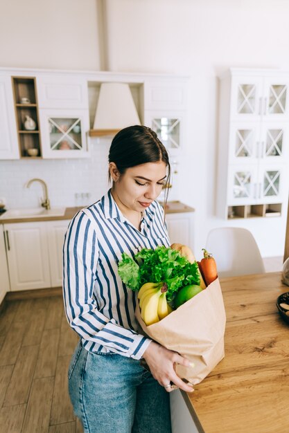 Jovem mulher caucasiana sorridente segura saco de compras eco com legumes frescos e baguete na cozinha moderna.