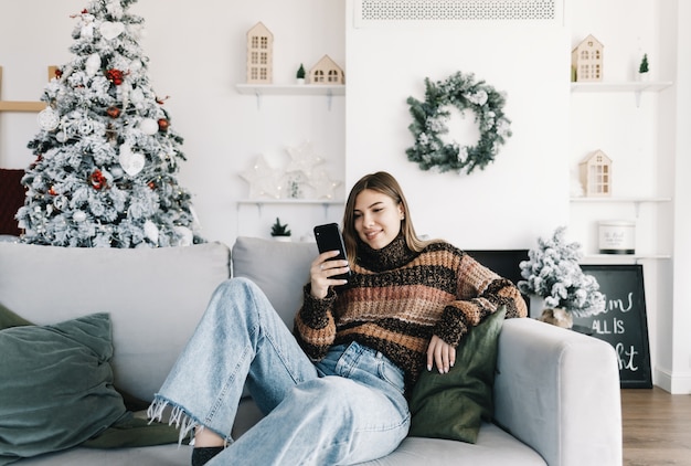 Jovem mulher caucasiana sorridente, usando telefone celular em férias em casa no sofá.